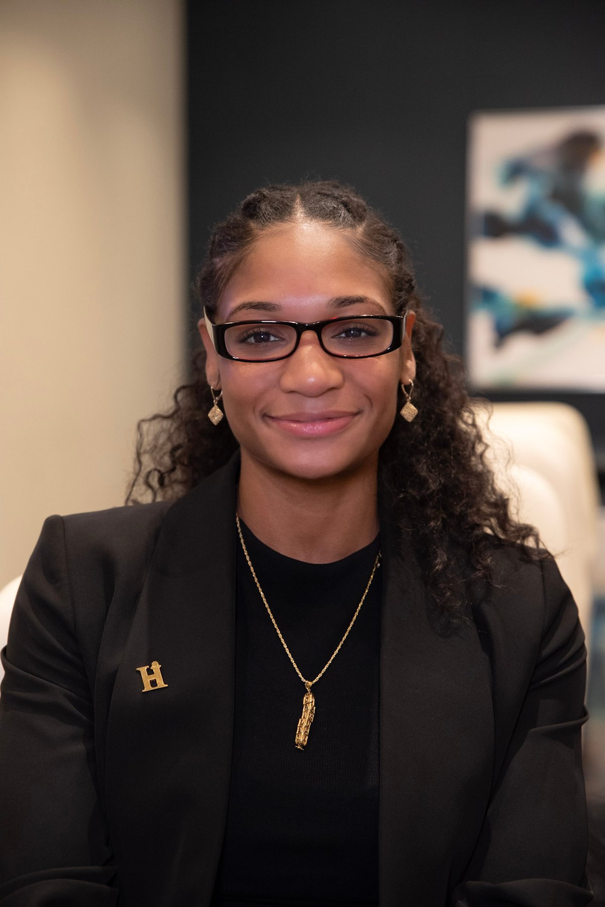 Professional headshot of Kaylan Julien wearing a black blazer, glasses, and gold jewelry against a blurred office background.